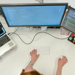 A female software engineer coding on dual monitors and a laptop in an office setting.