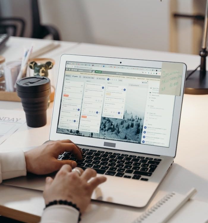 Hands typing on a laptop in a modern office setting with notes and task management software visible on the screen.
