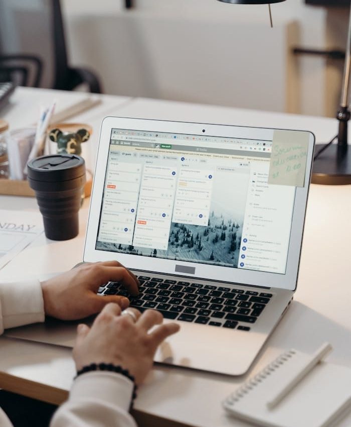 Hands typing on a laptop in a modern office setting with notes and task management software visible on the screen.