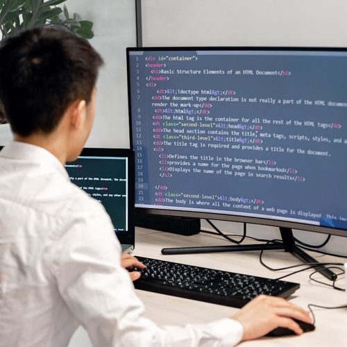 Adult male programmer coding on dual monitors in a modern indoor workspace.