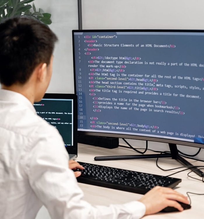 Adult male programmer coding on dual monitors in a modern indoor workspace.