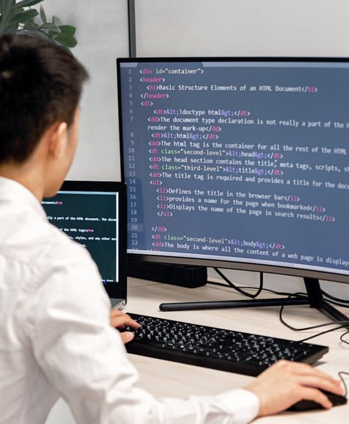 Adult male programmer coding on dual monitors in a modern indoor workspace.